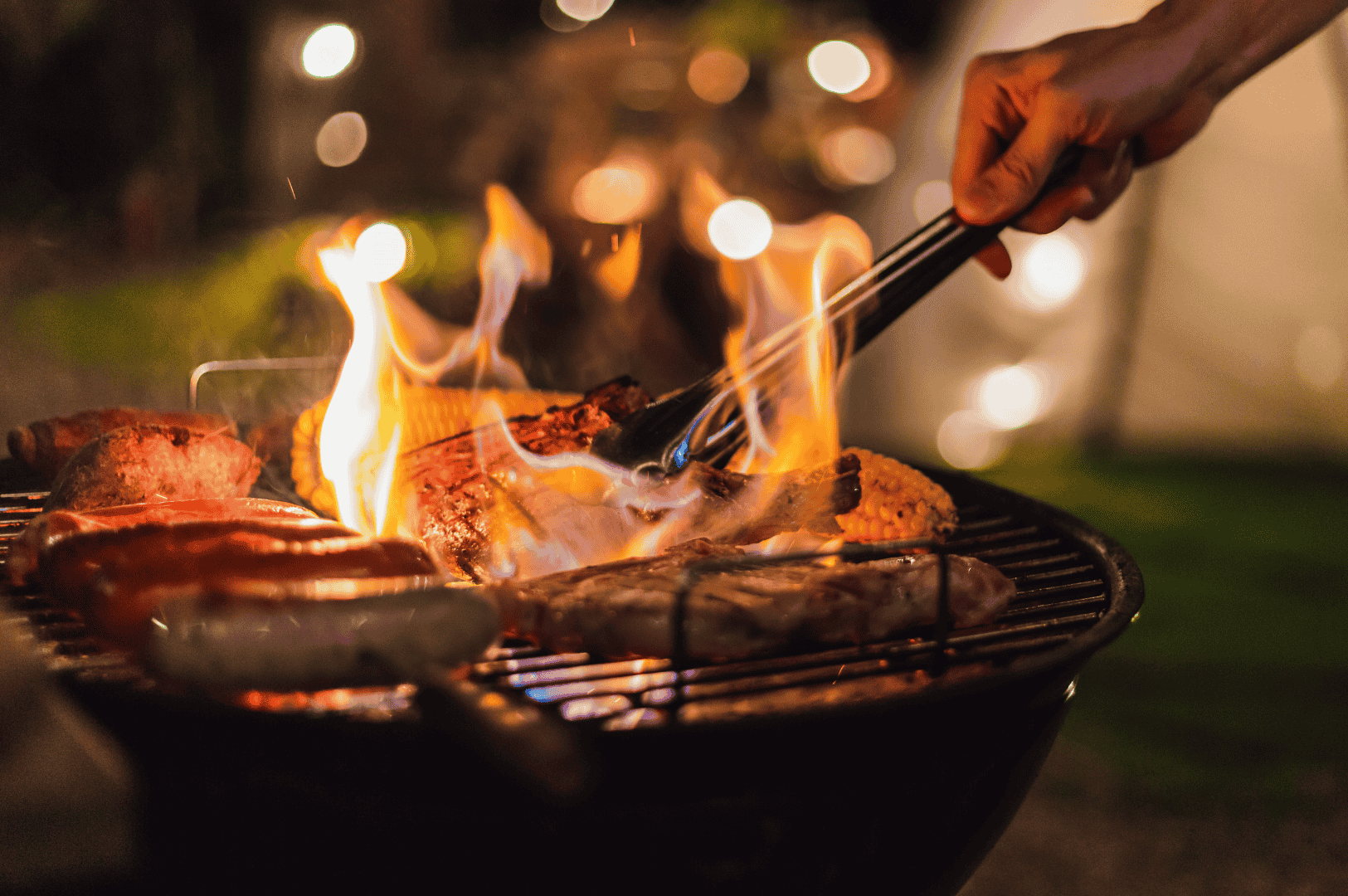 Close-up of a barbecue grill at dusk, with flames licking the edges of sizzling steaks and sausages. A hand with tongs adjusts the meat mid-cook, framed by glowing embers and soft bokeh in the background. The warm, directional lighting and shallow depth of field evoke the intimacy and drama of low-light food photography, capturing both the heat and human touch of outdoor grilling.
