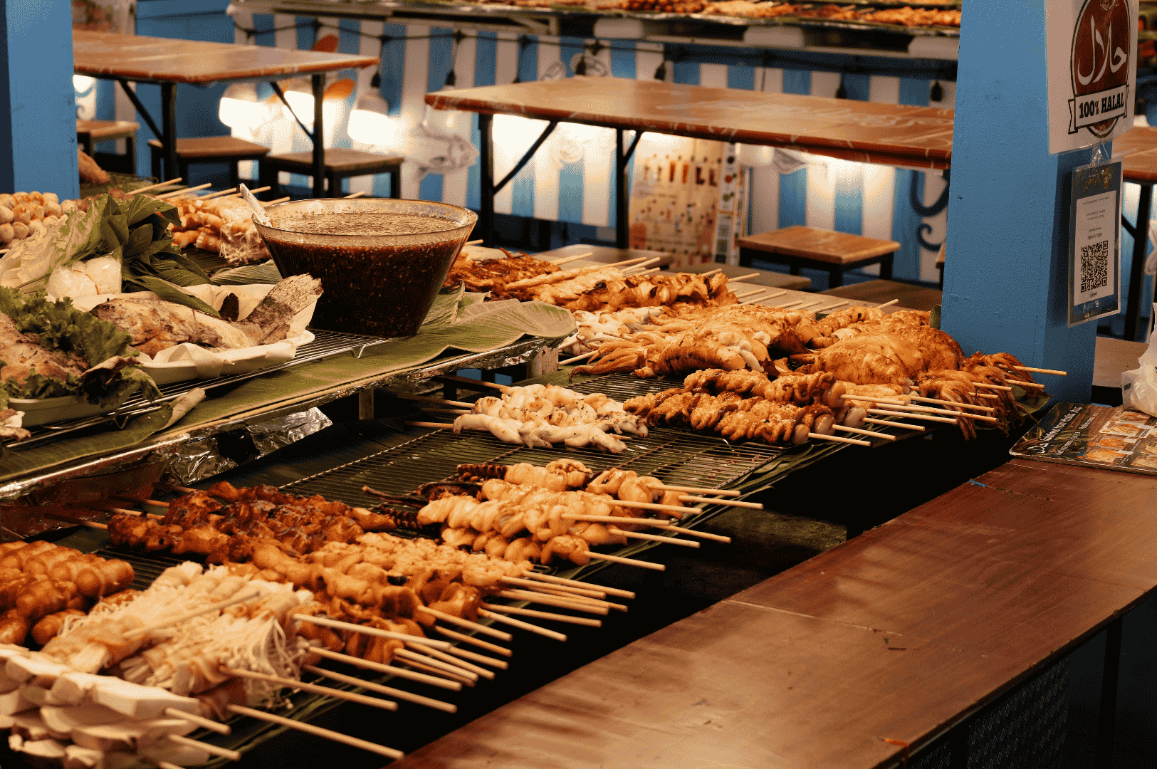Vibrant food stall scene showcasing rows of grilled skewers—chicken wings, mushrooms, seafood, and assorted meats—laid out on a gleaming metal surface. A bowl of sauce anchors the composition, while blue-and-white striped decor and dining tables in the background evoke a festive market atmosphere. The image is brightly lit, contrasting with previous low-light shots, offering a visual pivot in the editorial arc and highlighting how ambient lighting can shift mood and texture in food photography.