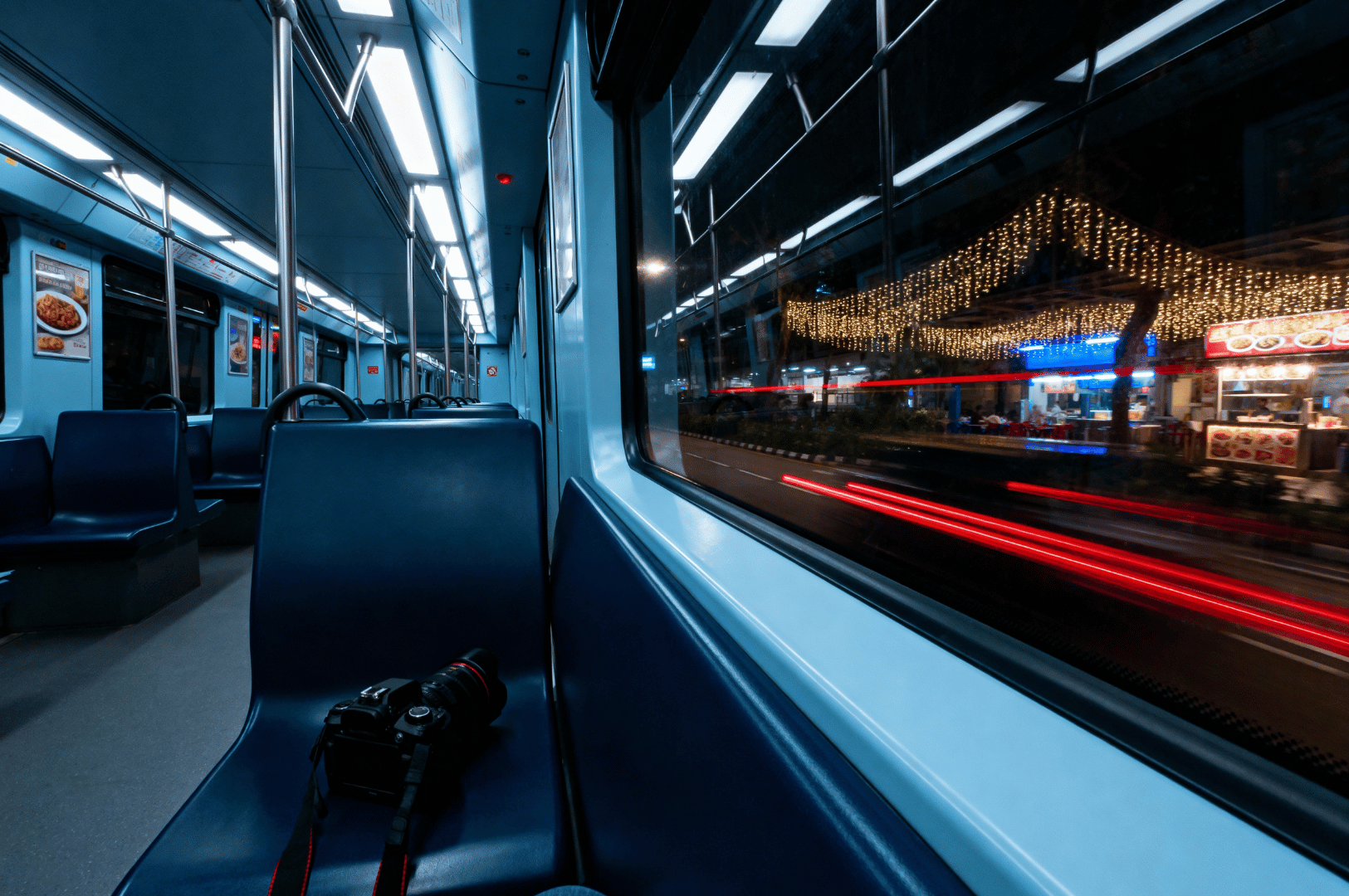 After-MRT- (1) Empty MRT car at night with a camera resting on a dark blue seat, contrasting the quiet interior with the glowing street outside, where festive lights and red vehicle trails hint at the city’s midnight energy—capturing the solitude and anticipation of a food photographer’s late-night journey.