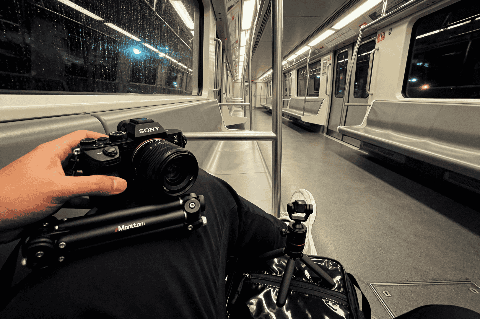 After-MRT- (3) Inside a dimly lit MRT car on a rainy night, a lone passenger in black pants and white sneakers balances a Sony camera and Manfrotto tripod on their lap, the raindrop-streaked windows framing the quiet intensity of a food photographer’s midnight journey.