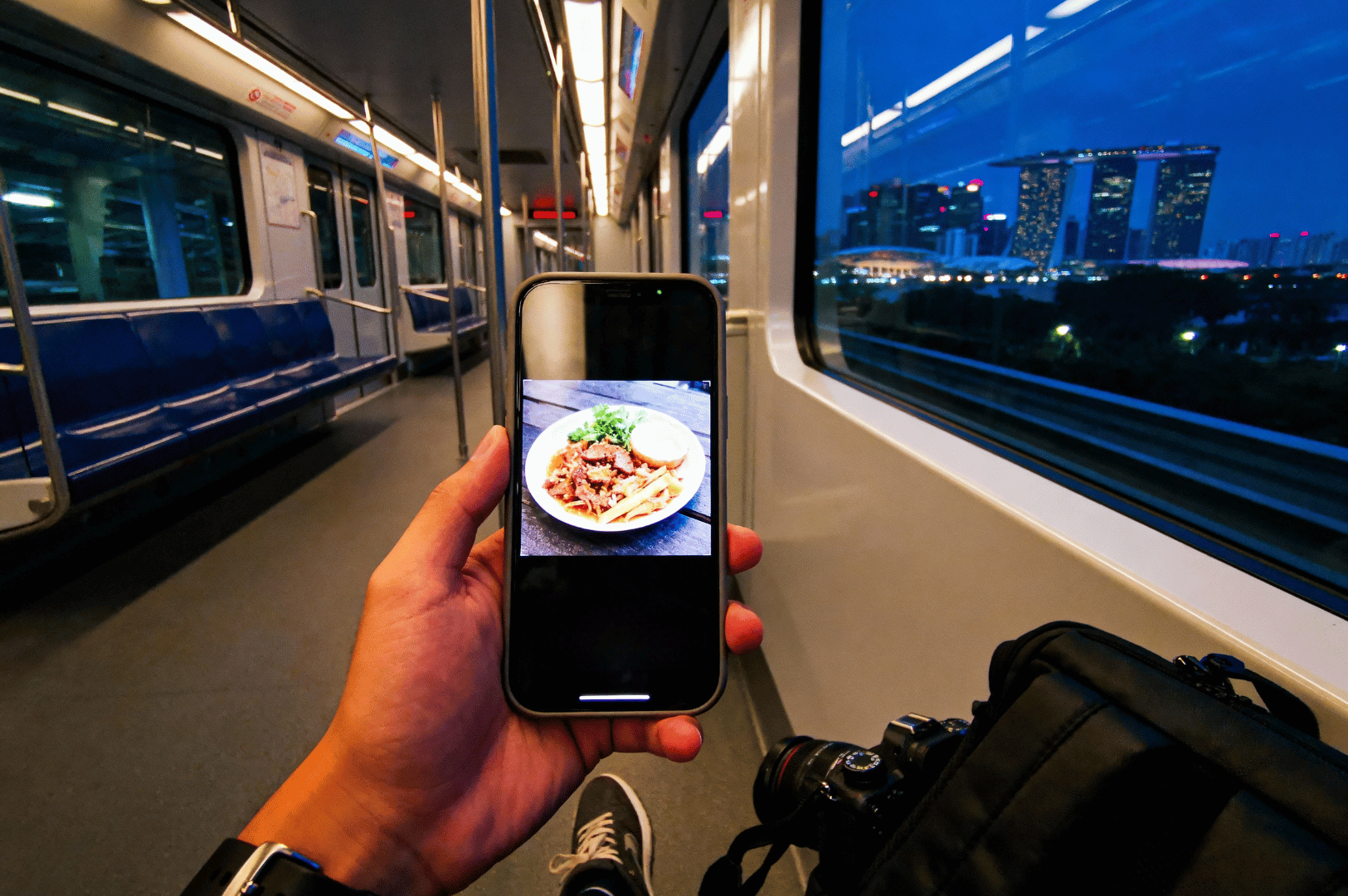 After-MRT- (5) Seated inside a twilight MRT car, a traveler holds a smartphone displaying a food photo while a camera bag rests nearby; through the window, Singapore’s Marina Bay Sands rises against the evening sky, blending the intimacy of culinary memory with the grandeur of urban exploration.