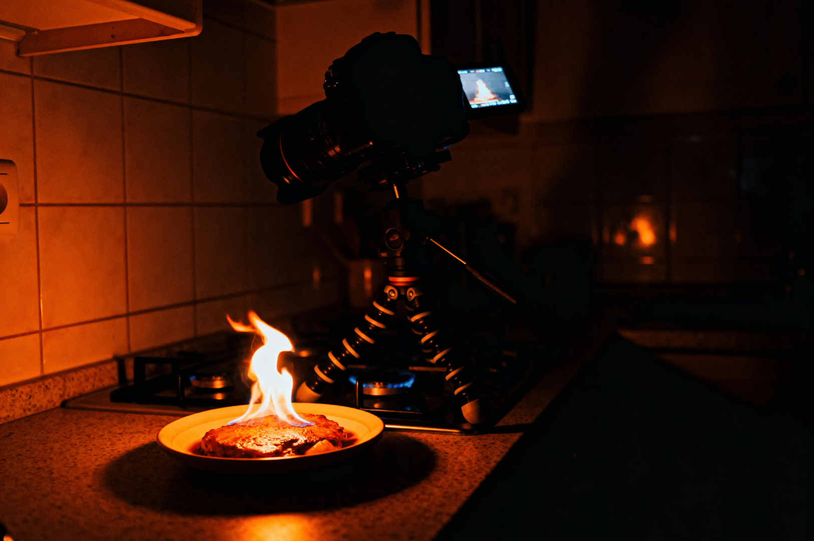 Flame-Photography- (3) A DSLR camera on a plate in a dim kitchen with a small flame rising from its top, illustrating creative light painting with household fire for night photography techniques.