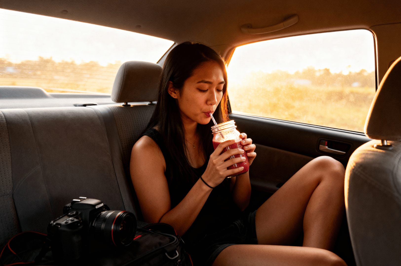 Food-Photographers-Diet- (4) Golden hour light spills into the backseat of a car where a food photographer sips from a mason jar, legs curled up in casual repose. A DSLR camera rests beside them on a soft bag—ready but momentarily forgotten. The scene captures a fleeting pause between shoots, where hydration and quiet reflection become part of the diet.