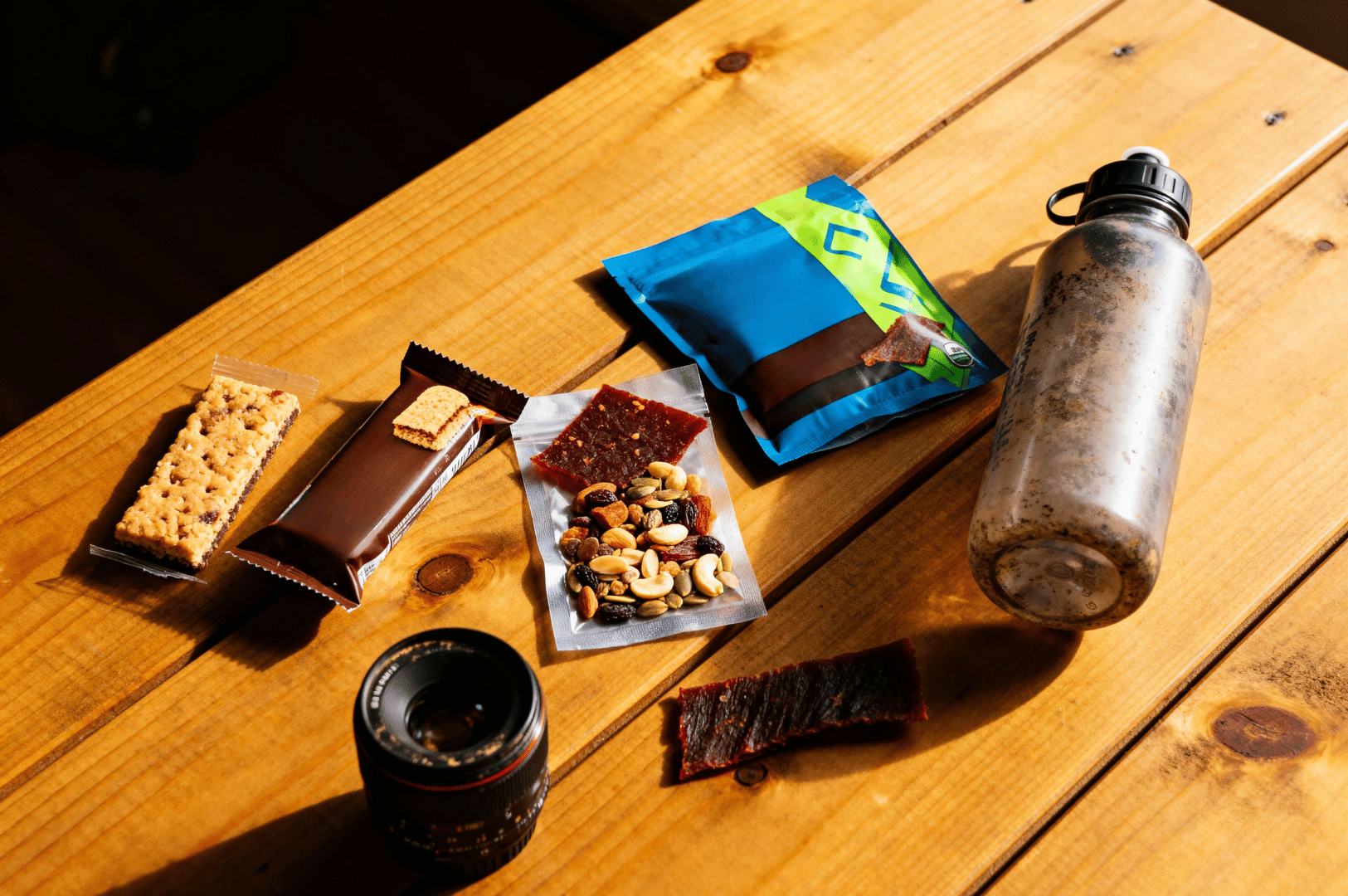 Food-Photographers-Diet- (5) Flat lay of a food photographer’s trail snacks: granola bars, chocolate-covered treats, jerky, trail mix, and a dirt-smudged water bottle—all arranged beside a camera lens on weathered wood. The scene captures the unglamorous reality of shoot-day sustenance, where nutrition meets necessity in the wild.