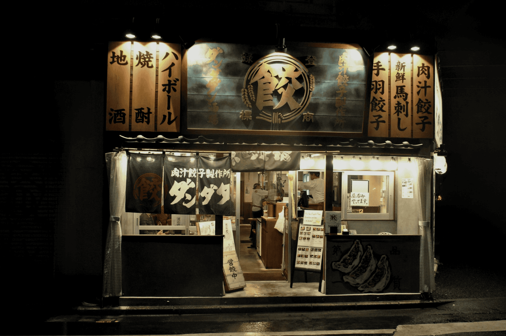 Tokyo-Late-Night-Ramen- (3) Warmly lit gyoza shop glowing against the night, with traditional wooden signage and handwritten menus promising juicy dumplings, grilled sake, and horse sashimi—an intimate glimpse into Tokyo’s late-night food rituals and secret flavors.