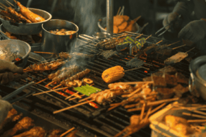 Lowlight-Food-Photography-1 Street-side barbecue scene captured in low light, with glowing embers illuminating skewers of meat, seafood, and vegetables. Smoke curls upward as gloved hands tend the grill, creating dynamic motion and texture. The warm tones and contrast highlight the communal energy and rich detail of open-flame cooking, showcasing the power of ambient light and shadow in food photography.