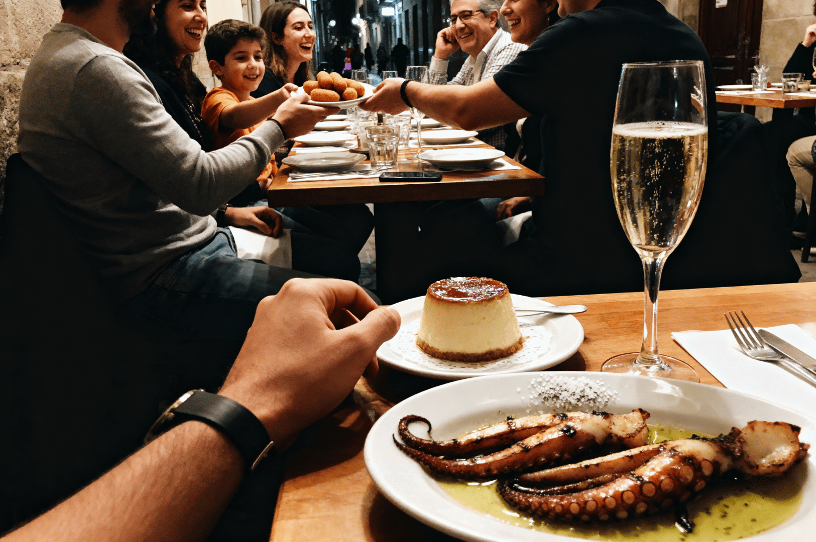Festive late-night dinner in a lively Barcelona restaurant, captured from a diner’s perspective. A plate of grilled octopus drizzled with olive oil and herbs, a creamy flan dessert, and a glass of sparkling wine anchor the foreground, while friends smile and converse around the table. The warmly lit backdrop of fellow diners and vibrant ambiance evokes the joy and intimacy of hidden culinary gatherings after dark.