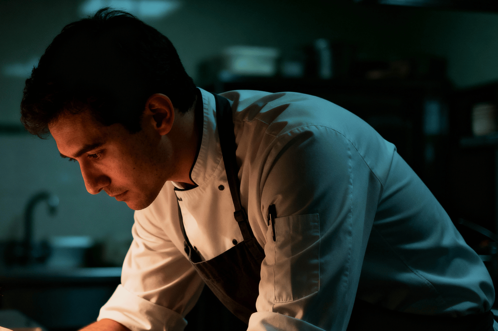 Chef-Portrait- (5) Focused chef in a dim kitchen, leaning over a plate as warm light highlights his face and white jacket.