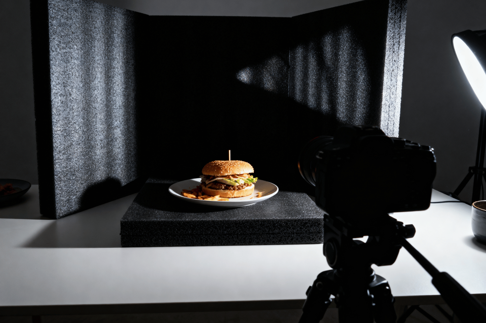 A studio light casts crisp highlights on a burger and fries, framed by black foam boards and a camera tripod—revealing the precision and contrast-driven setup behind dramatic food photography.