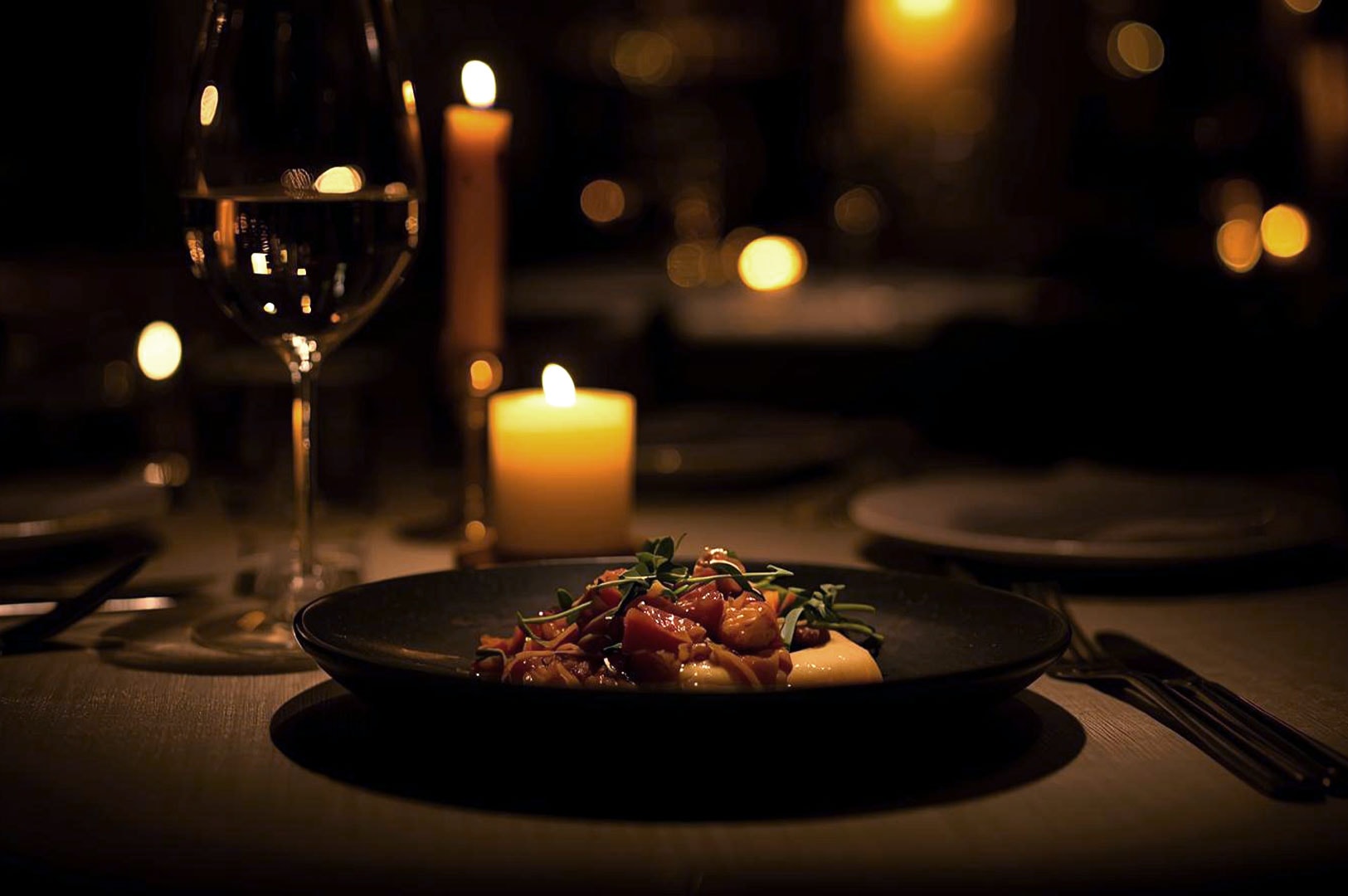 A romantic, dimly lit table with a glass of white wine, two lit candles, and a gourmet dish plated on a dark surface.