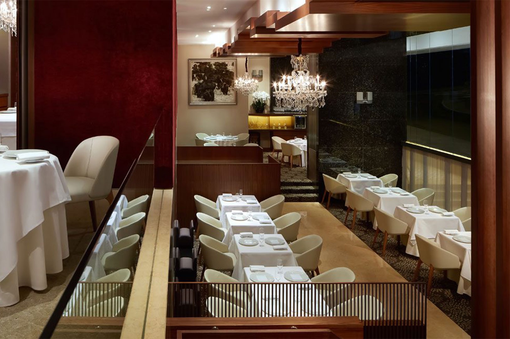 A spacious, upscale restaurant interior featuring crystal chandeliers, dark wood accents, and rows of dining tables with white linens and cream chairs.