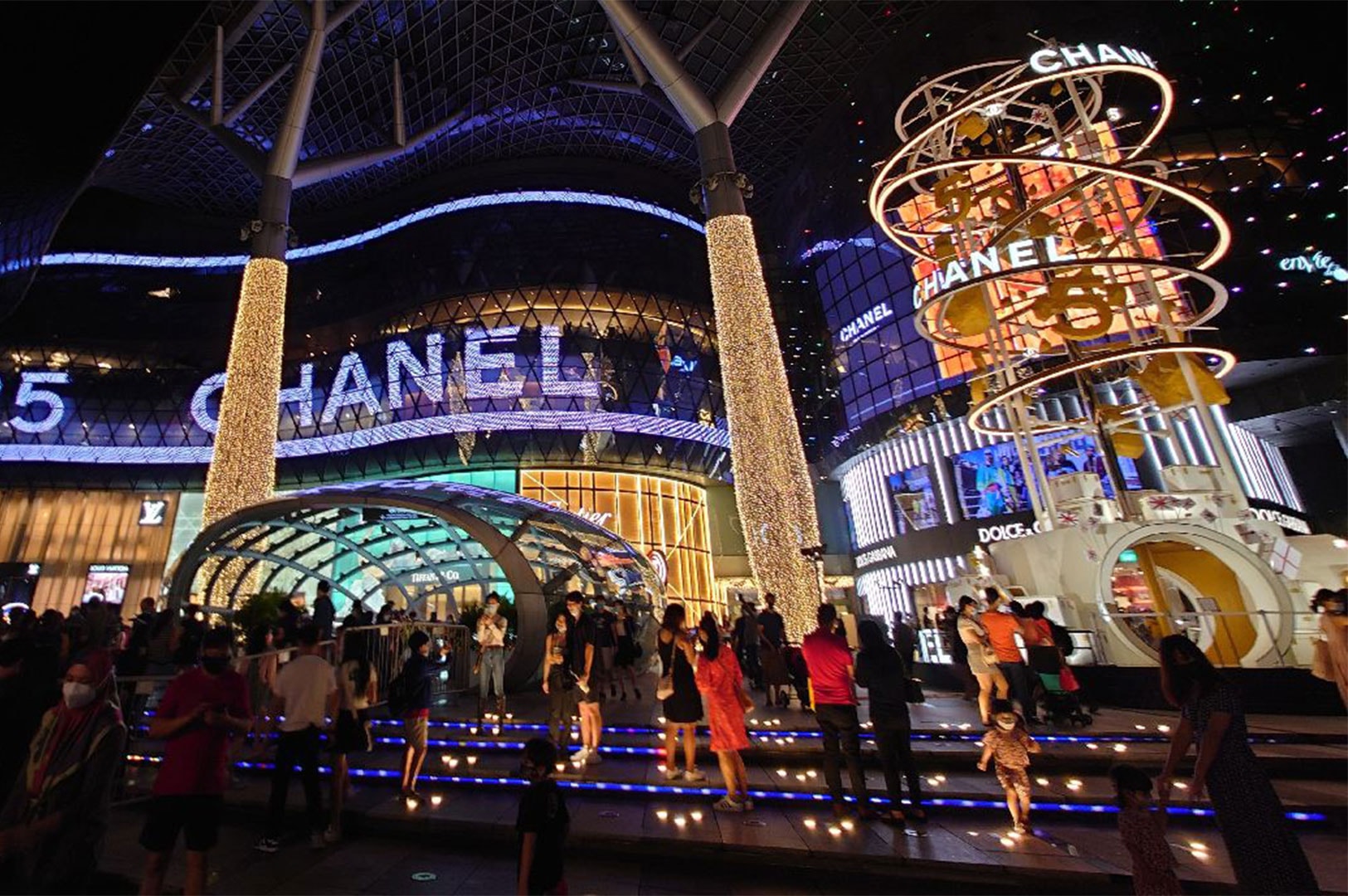 Crowded street scene in Singapore at night, showing shoppers gathered near the brightly lit Christmas decorations and retail displays, including a large 'Chanel' sign.