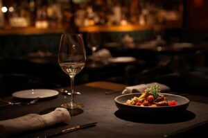 Close-up shot of a wine glass and a plated dish with vegetables and meat, set on a dark table in a dimly lit, upscale restaurant.