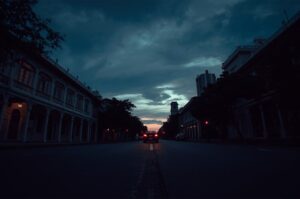A moody urban street scene at twilight featuring colonial-style architecture and a car driving away with red taillights glowing against the blue sky.