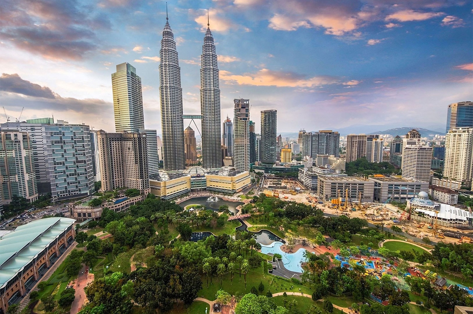 An aerial landscape view of the Kuala Lumpur city skyline at sunset, prominently featuring the Petronas Twin Towers and the KLCC park below.