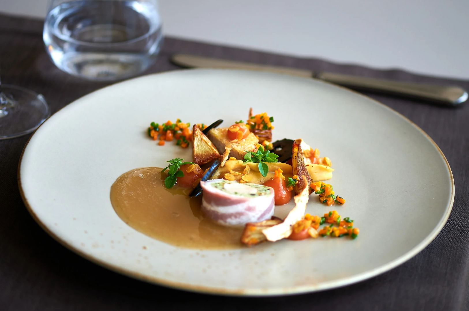 Best-Dining-2 Gourmet dish on a white plate featuring a bacon-wrapped roulade, colorful purees, diced vegetables, and artistic sauce presentation, exemplifying fine dining elegance.