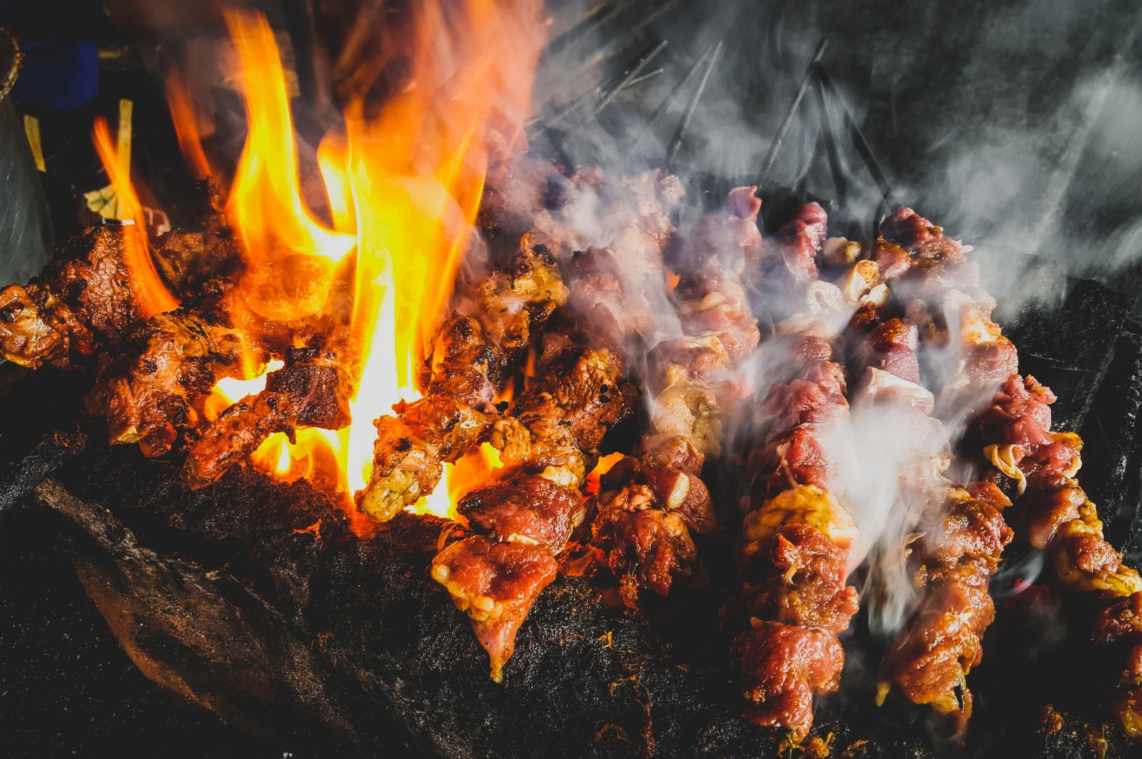 Best-Dining-3 Skewers of marinated meat grilling over open flames on a charcoal grill, with smoke and fire rising, capturing the intensity and flavor of street-style cooking.