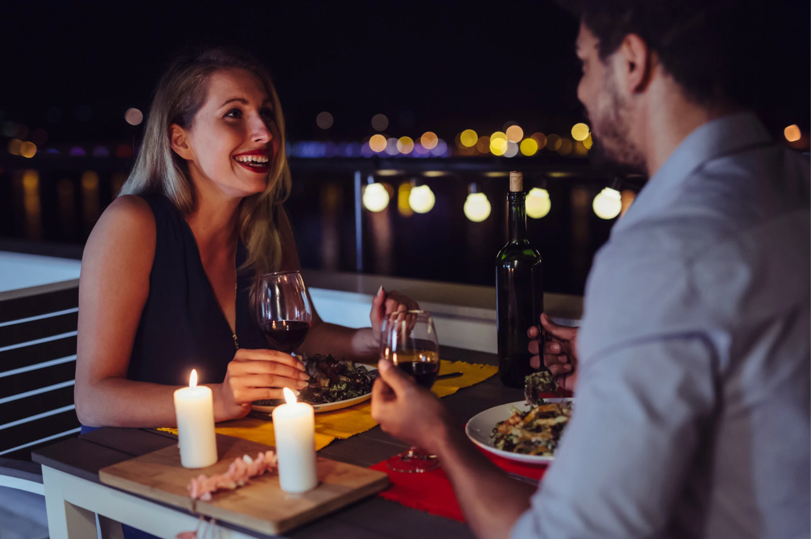 Best-Dining-4 Two people enjoying a candlelit dinner with wine on a balcony at night, overlooking city lights reflected on water, creating a romantic and intimate atmosphere.