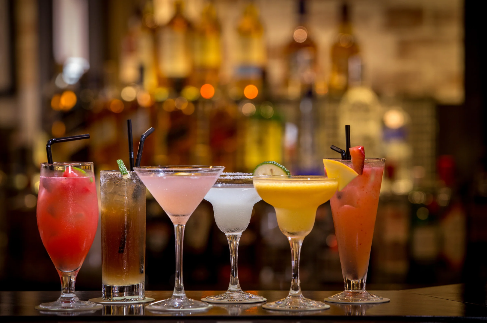 Best-Dining-5 Six colorful cocktails lined up on a bar counter, each uniquely garnished and served in different glassware, with liquor bottles blurred in the background.