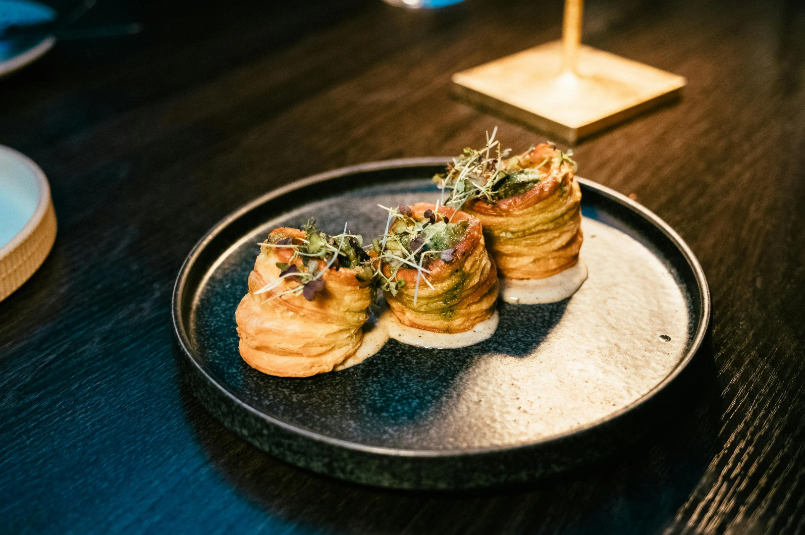 Best-Dining-6 Three stacked potato spirals garnished with microgreens on a dark ceramic plate, each resting on a pool of creamy white sauce, showcasing refined plating in a fine dining setting.