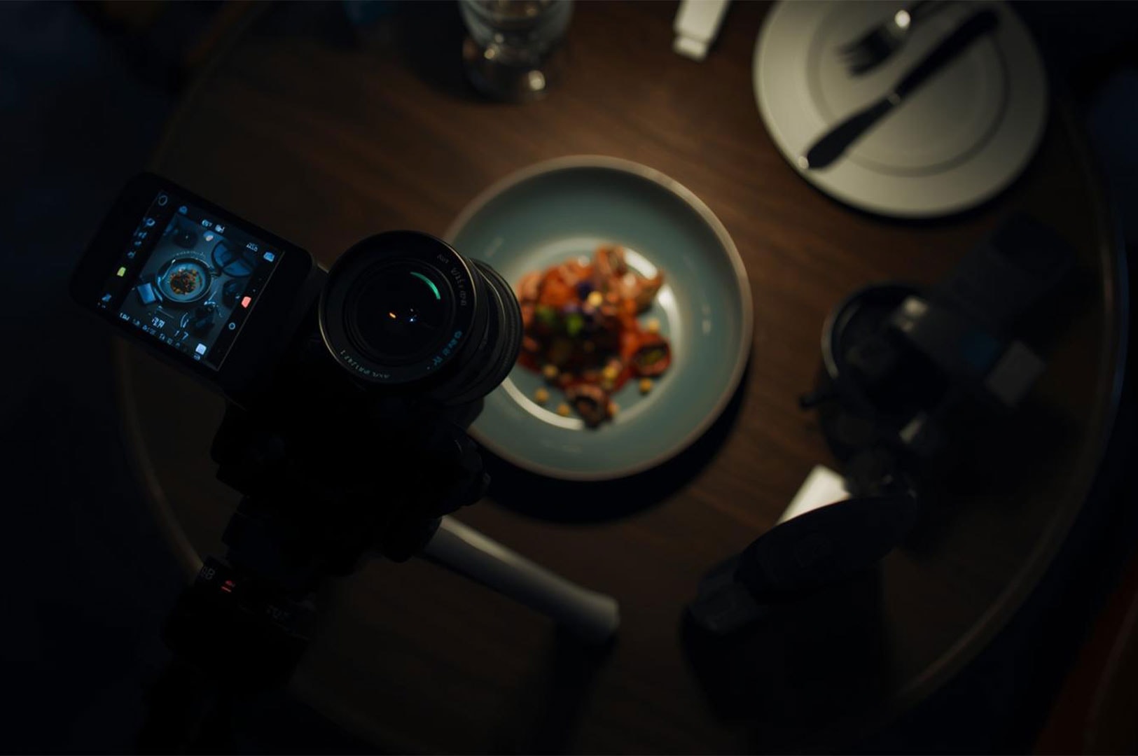 Overhead, low-light shot showing a camera on a tripod taking a picture of a dish in a blue bowl on a dark wooden restaurant table, demonstrating food photography.