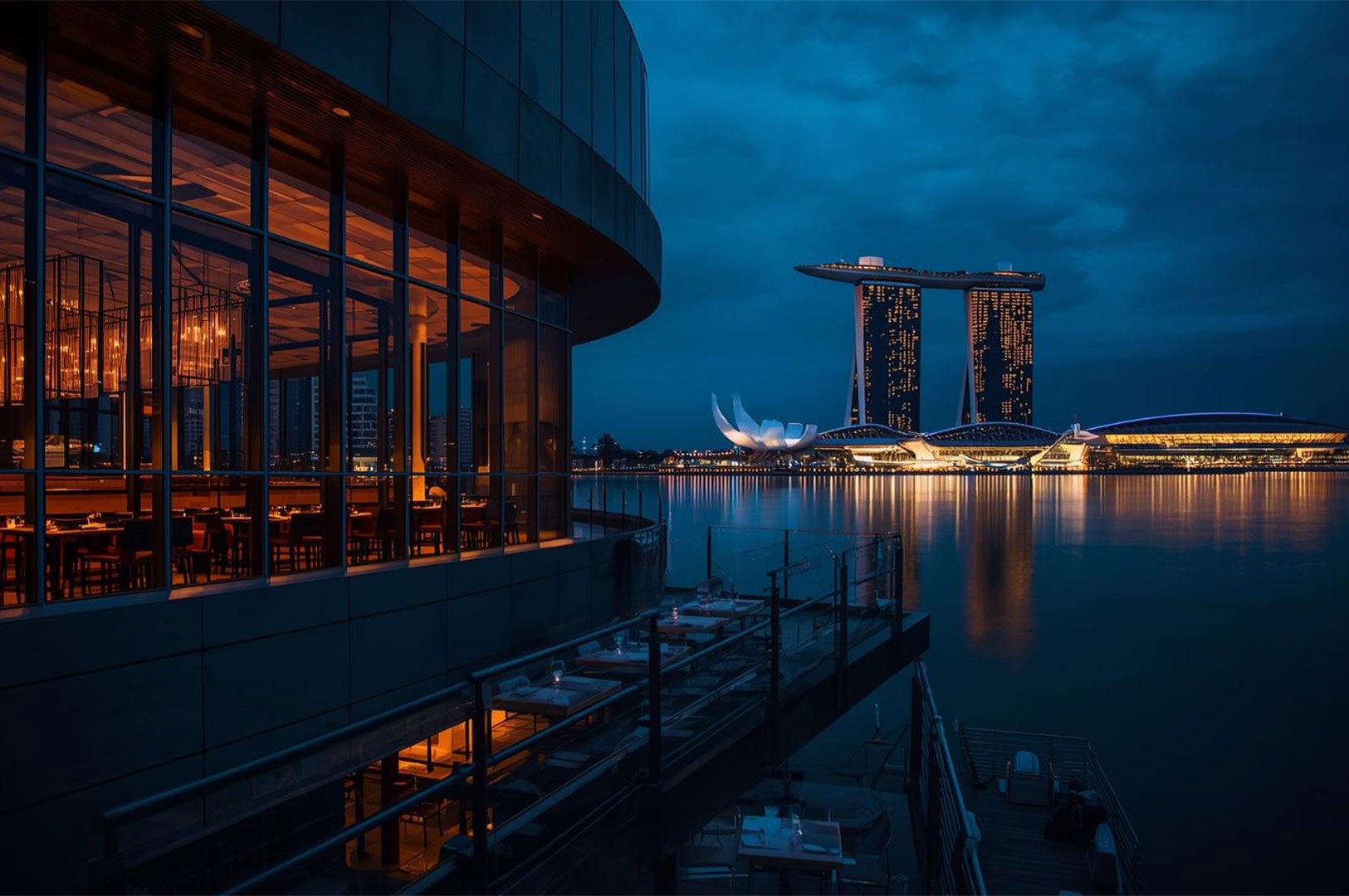 View of a modern, glass-fronted waterfront restaurant in Singapore at dusk, overlooking Marina Bay and the illuminated Marina Bay Sands hotel.