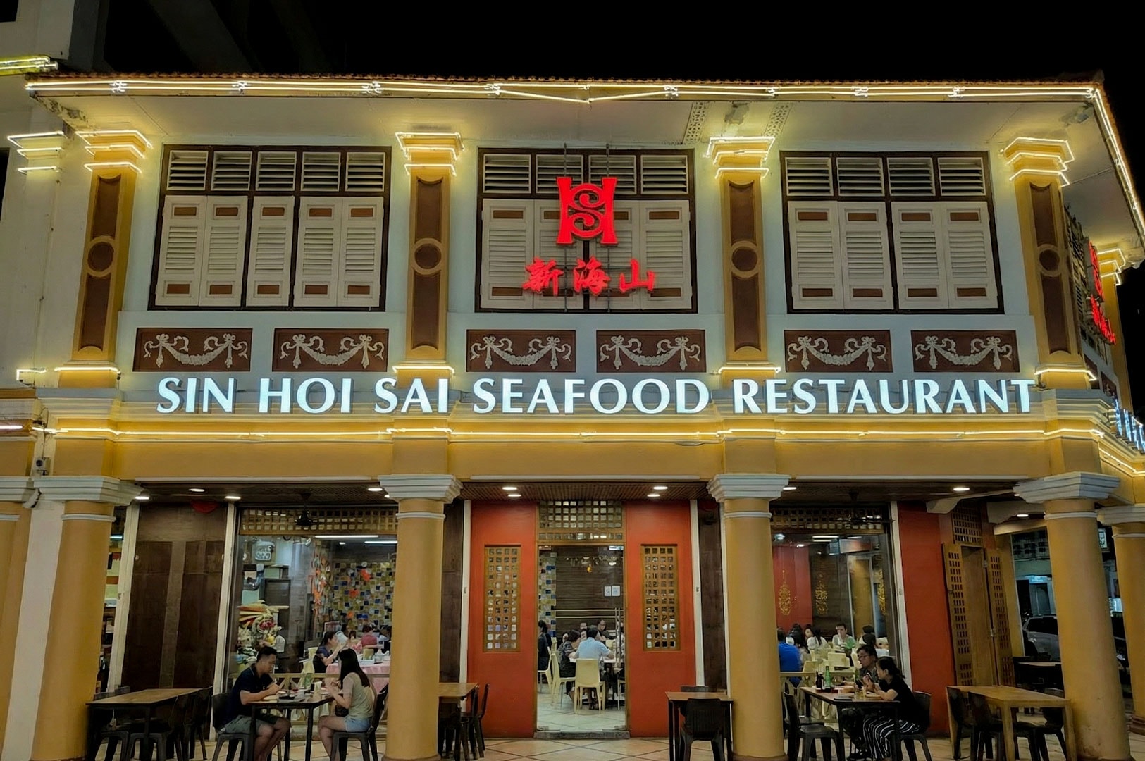 The illuminated exterior of Sin Hoi Sai Seafood Restaurant at night, showcasing traditional shophouse architecture and diners sitting at tables outside.