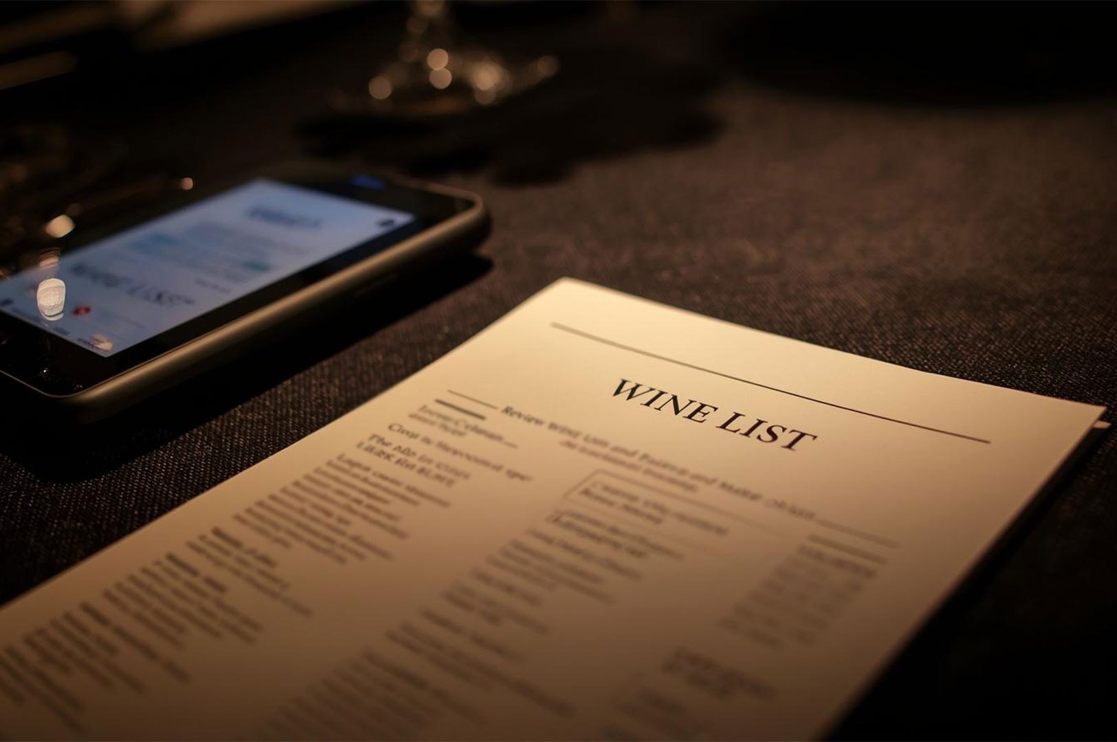A close-up of a printed "WINE LIST" menu and a smartphone resting on a dark table in a restaurant setting.