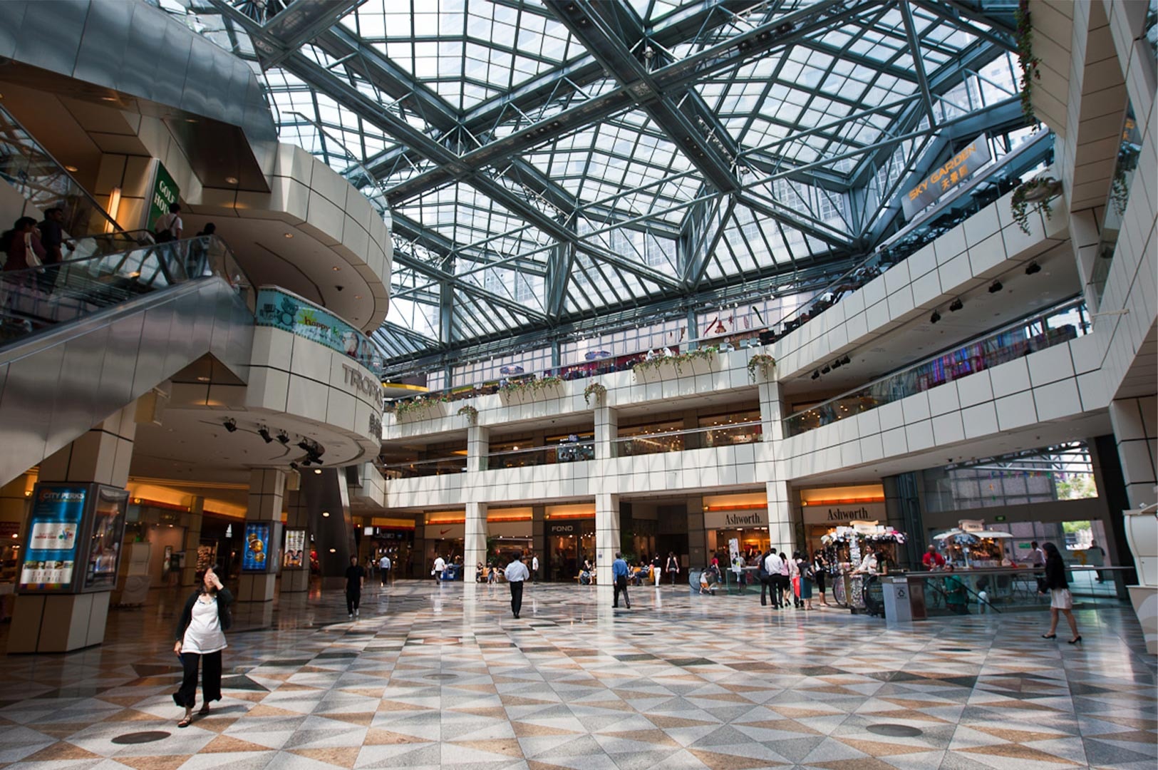 Interior of a multi-level shopping mall featuring a massive geometric glass atrium roof, tiled flooring, and shoppers walking near storefronts and escalators.