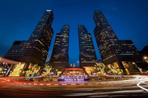 Low angle night view of the Suntec City towers and Fountain of Wealth in Singapore, featuring long-exposure light trails from passing traffic.