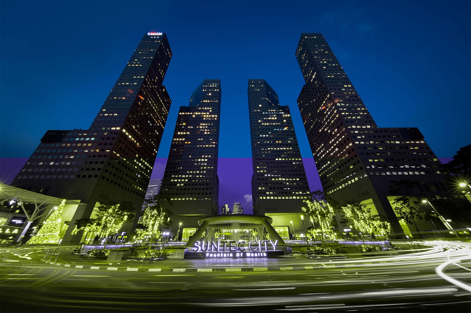 Low angle night view of the Suntec City towers and Fountain of Wealth in Singapore, featuring long-exposure light trails from passing traffic.