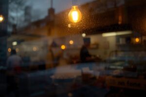 A close-up of a glowing filament lightbulb hanging against a rain-splattered window, with a blurry, bokeh view of a city street in the background.