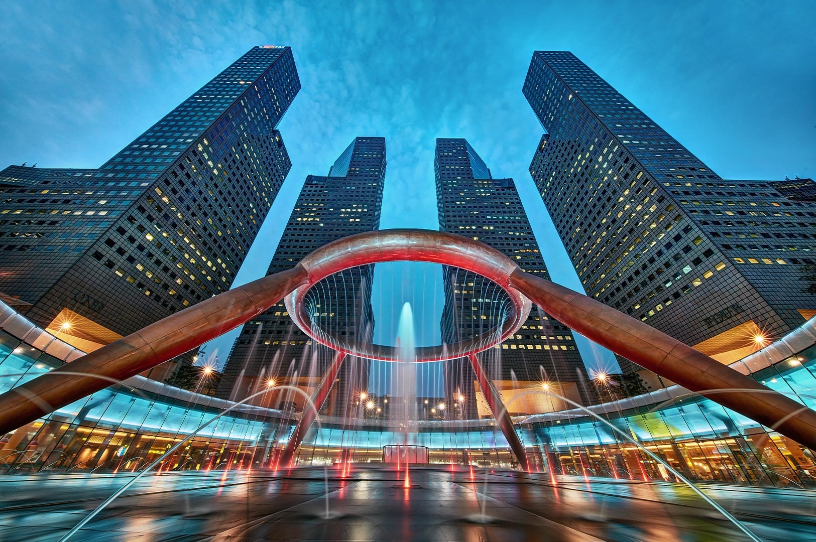 Low-angle view of the Fountain of Wealth in Suntec City, Singapore, illuminated at night with tall skyscrapers towering in the background.