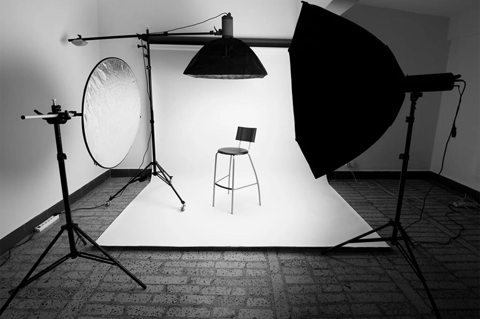 Black and white photograph of a professional studio setup featuring a high stool, boom arm strobe light, and softbox reflector against a white backdrop.