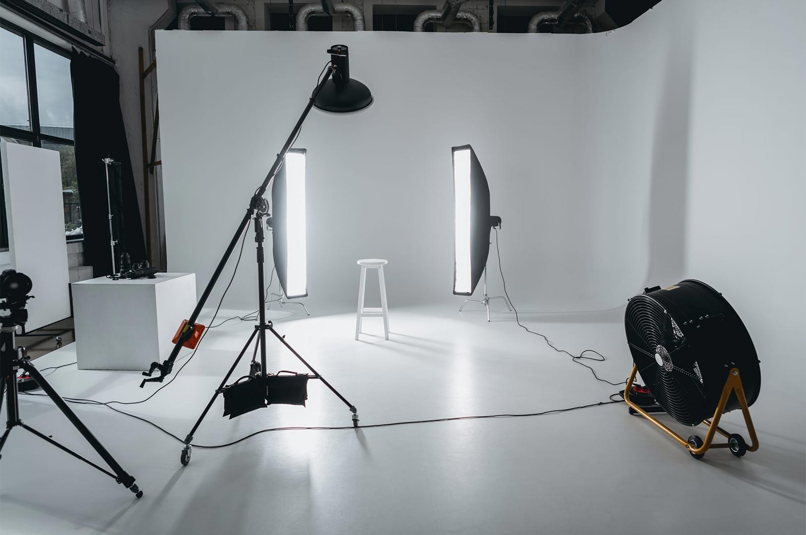 Behind-the-scenes view of a spacious photo studio with a white cyclorama wall, featuring strip box lights, a heavy-duty floor fan, and a camera mounted on a tripod.