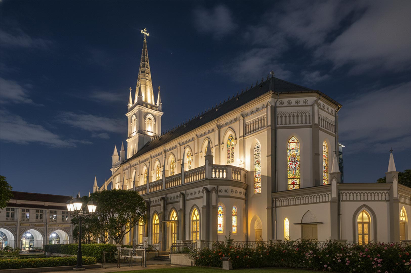 Exterior night view of the historic CHIJMES Hall in Singapore, featuring its illuminated Gothic Revival architecture, spire, and stained glass windows under a twilight sky.