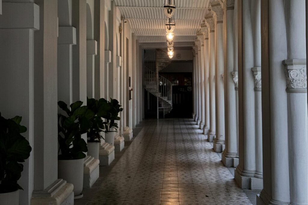 A long, dimly lit historic hallway with white Doric columns, decorative floor tiles, and a spiral staircase in the background.