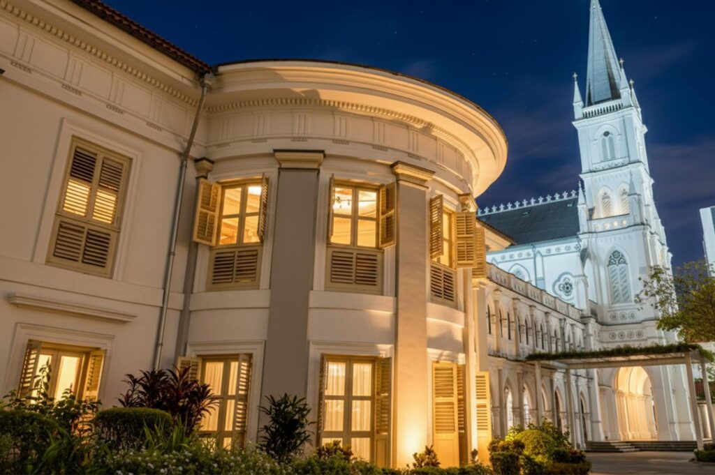 Close-up exterior of the CHIJMES heritage building with warm interior lighting glowing through shutters, with the white chapel spire visible in the background.