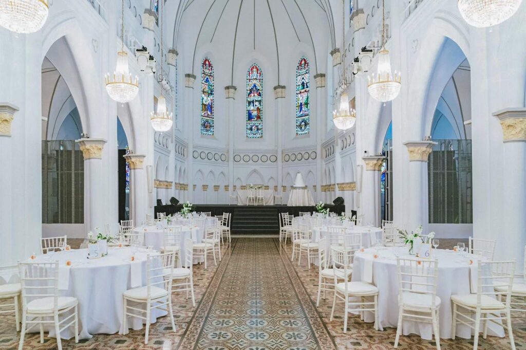 A wedding reception setup inside CHIJMES Hall, featuring round tables with white tablecloths, white Tiffany chairs, and a stage positioned beneath stained glass windows.
