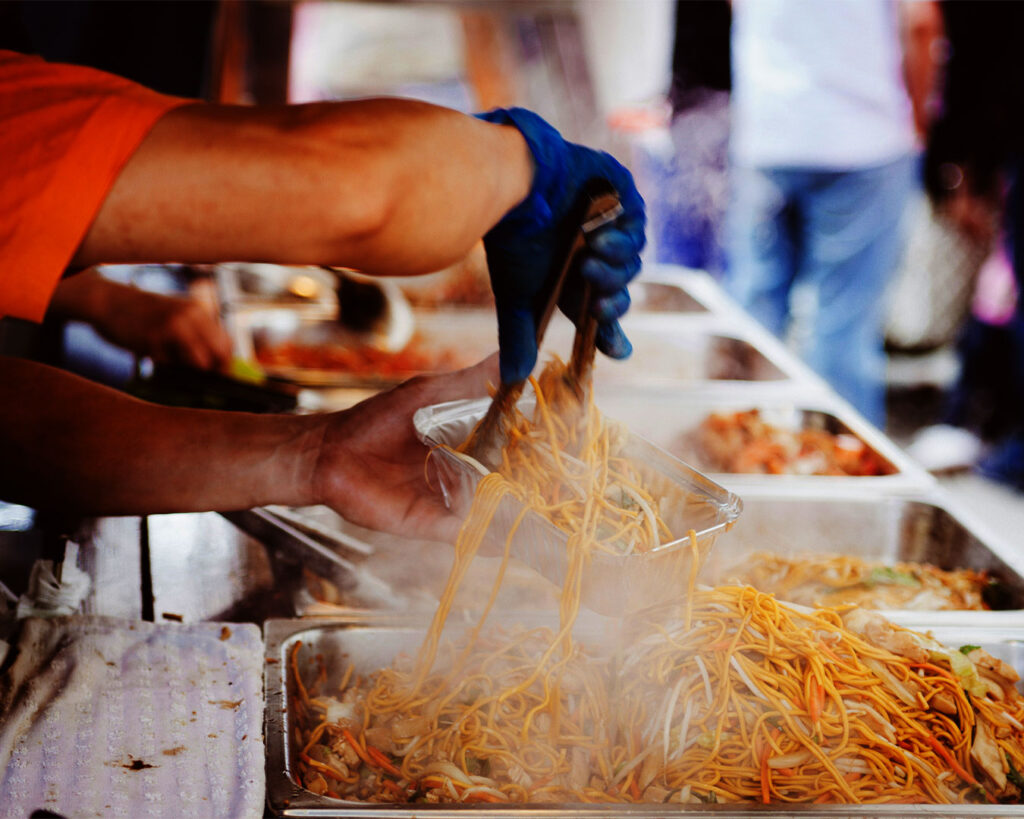 A pair of hands packing freshly made spring noodles into a container, showing the noodles’ soft texture and light flour dusting in a small kitchen or stall workspace.