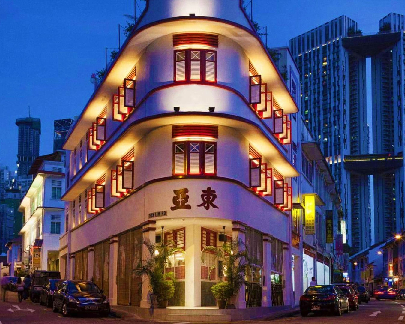A night view of Tanjong Pagar street illuminated by warm shop lights and street lamps, with restaurants and bars lining both sides of the road and pedestrians walking through the lively evening atmosphere.