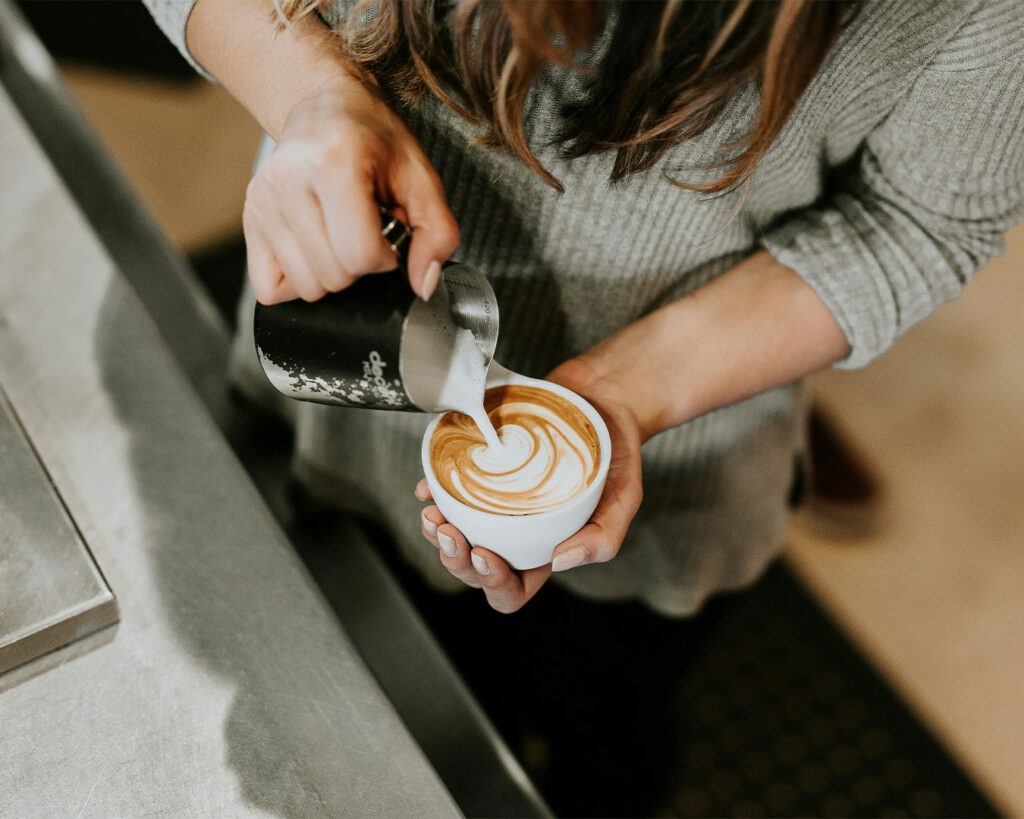 A barista carefully pouring steamed milk into a cup of coffee, forming latte art on the surface, with focus on the flowing milk stream and the smooth crema creating a patterned design.