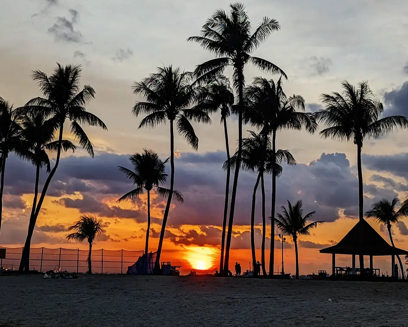 Tanjong Beach Club at sunset, with the sky painted in warm orange and pink hues reflecting over the calm beach and pool area, creating a romantic evening atmosphere.