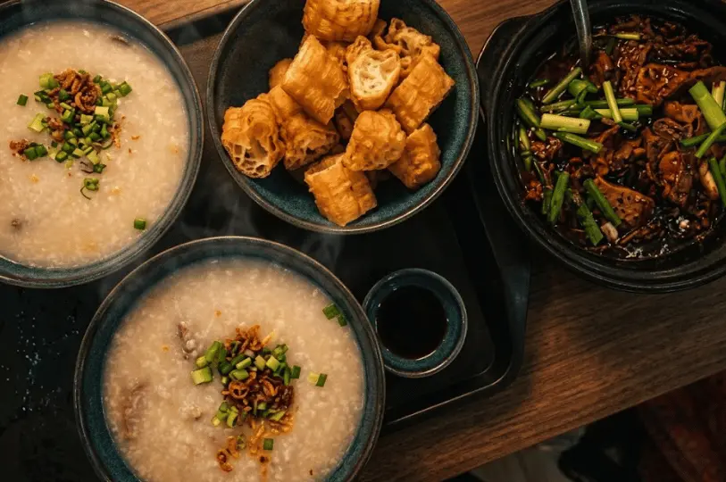 Top-down composition of a comforting late-night supper tray in Geylang, featuring bowls of warm congee, crispy youtiao, and dark braised dishes captured with warm, low-key lighting.
