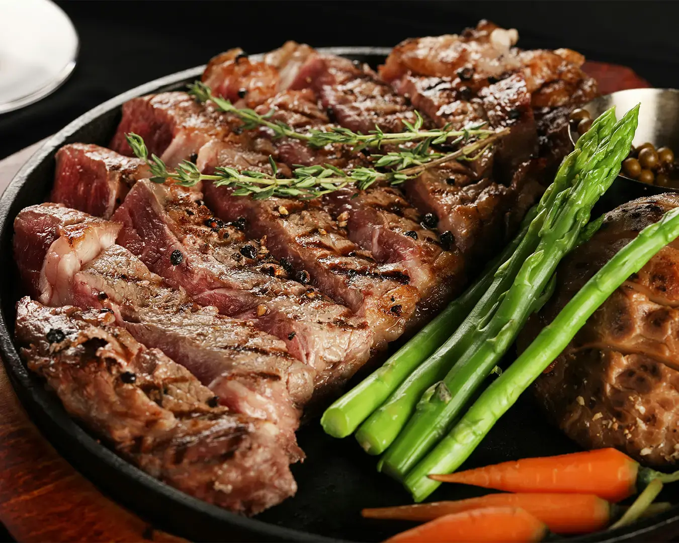 Grilled steak slices garnished with herbs, accompanied by asparagus and baby carrots on a sizzling platter, exuding a savory and appetizing aroma.
