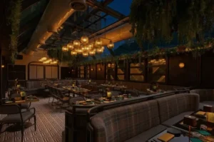 Upscale restaurant interior at night with a glass ceiling revealing the dark sky, hanging greenery, a large glowing chandelier, and neatly set dining tables with plaid booth seating.