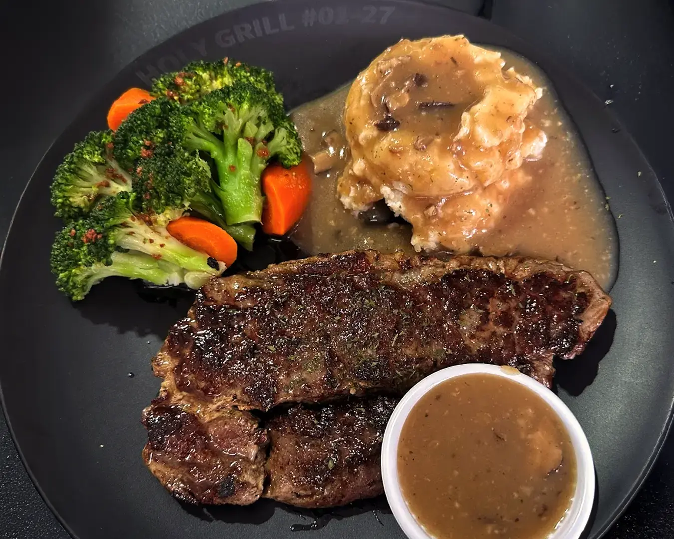 Grilled steak with a rich brown crust, mashed potatoes with gravy, and steamed broccoli and carrots on a black plate, evoking a hearty meal.