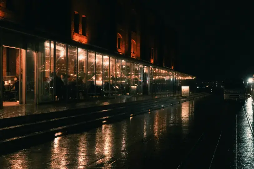 A nighttime exterior view of a lively restaurant housed in a brick building with a long, illuminated glass facade, casting warm light reflections onto the wet pavement outside.