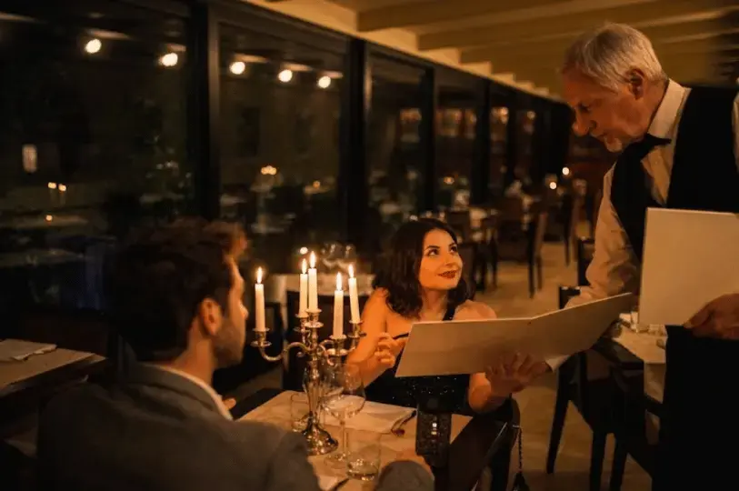 A waiter in a black vest presents a menu to a smiling woman sitting across from a man at a romantic, candlelit restaurant table beside a dark window.