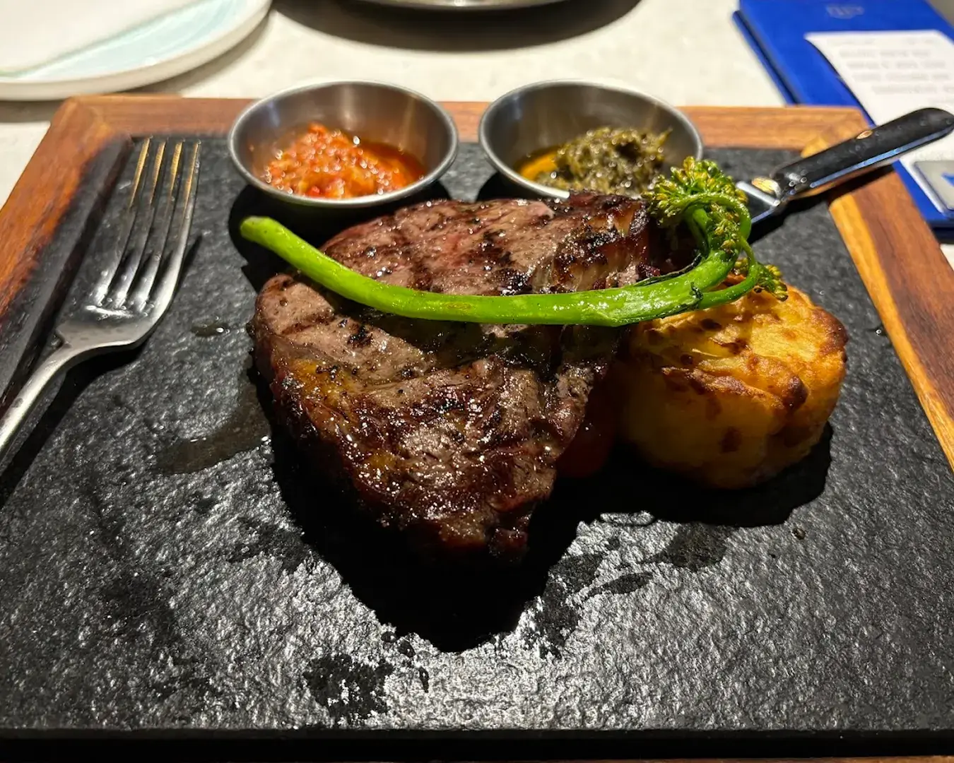 Grilled steak on a black slate plate, garnished with a broccolini stalk. Served with two small metal bowls of sauces and a baked potato stack.